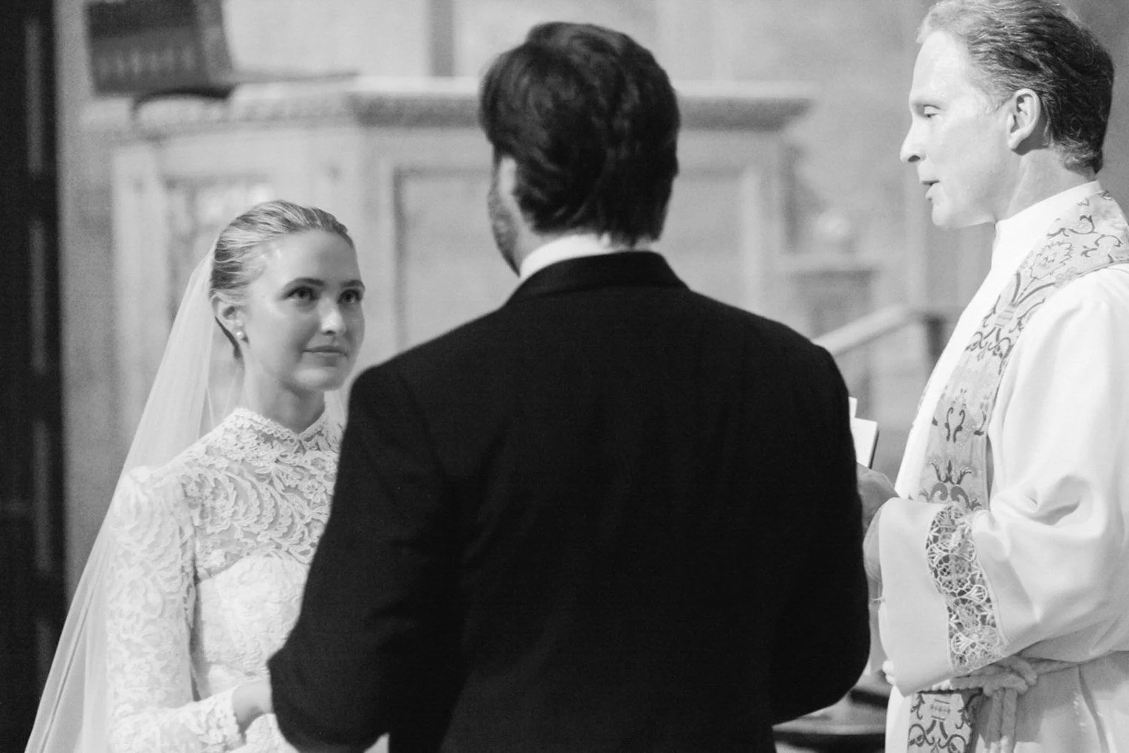 A black and white scene of a wedding ceremony inside a church. The bride, with her hair in an updo and wearing a laced wedding dress, is looking at the groom. The groom, seen from the back, is wearing a dark suit. The officiant, dressed in religious 