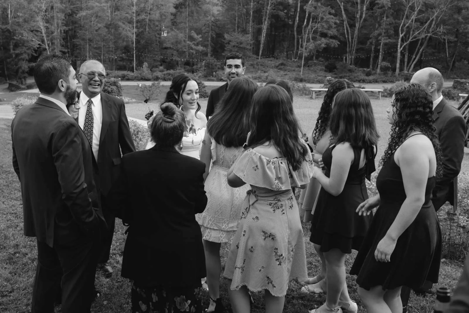 A group of people dressed in formal attire gathered outdoors on a lawn, engaged in conversation, with trees and a pathway in the background.