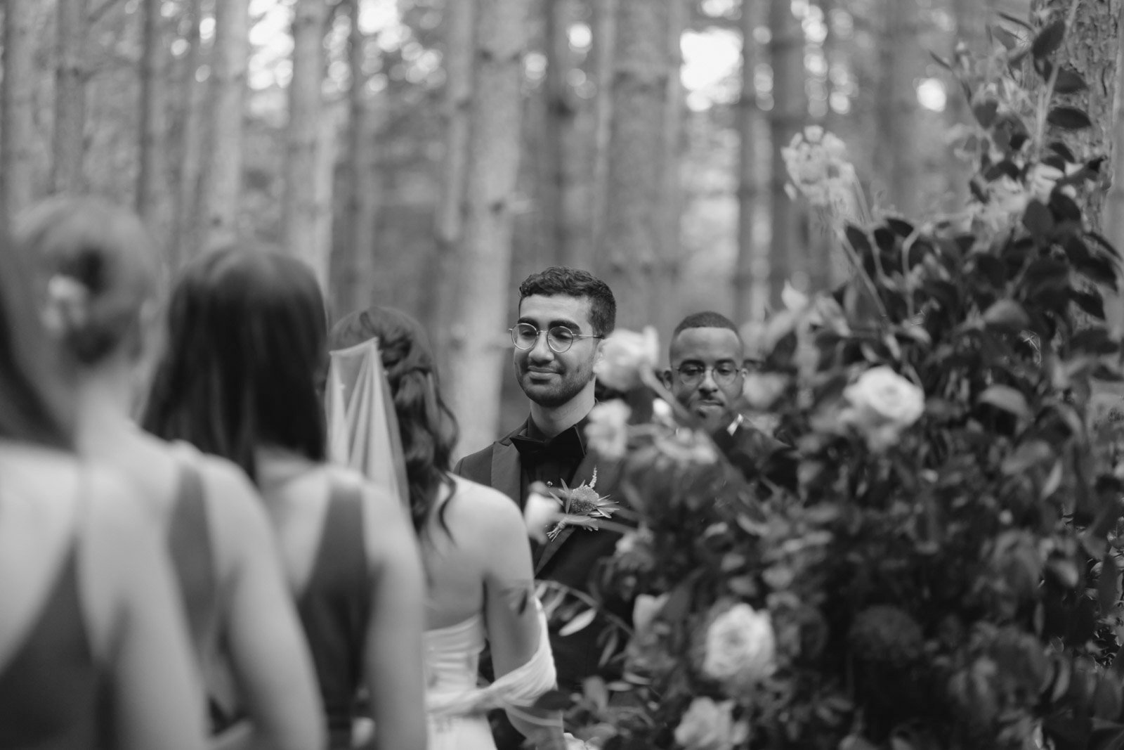 A black and white photo of a wedding ceremony in a forest, with a group of people including the bride and groom, facing each other, surrounded by trees and floral arrangements.