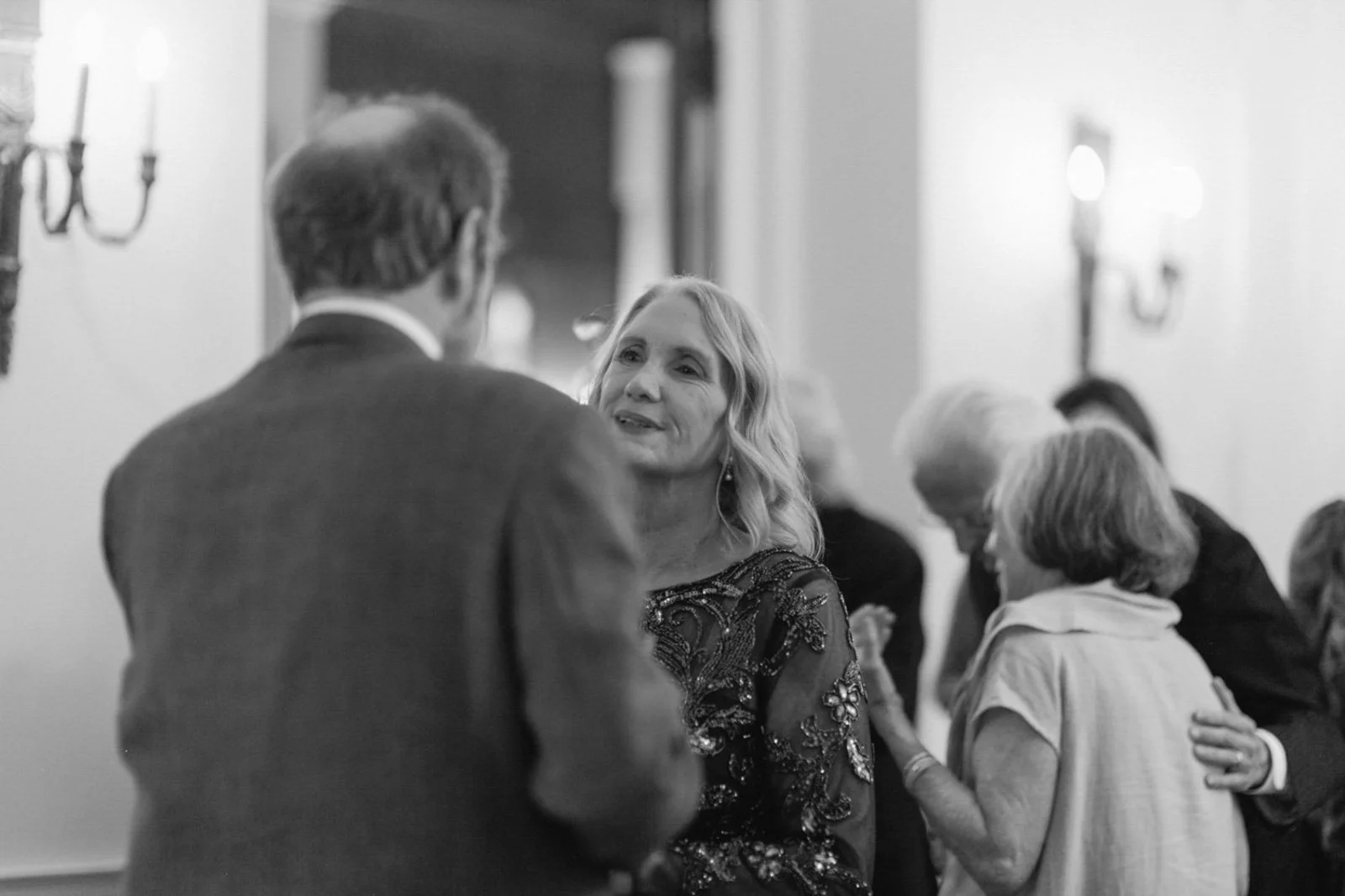 People socializing at a formal event, including a woman with wavy hair wearing a decorative dress. The scene appears indoors with wall sconces and blurred background.
