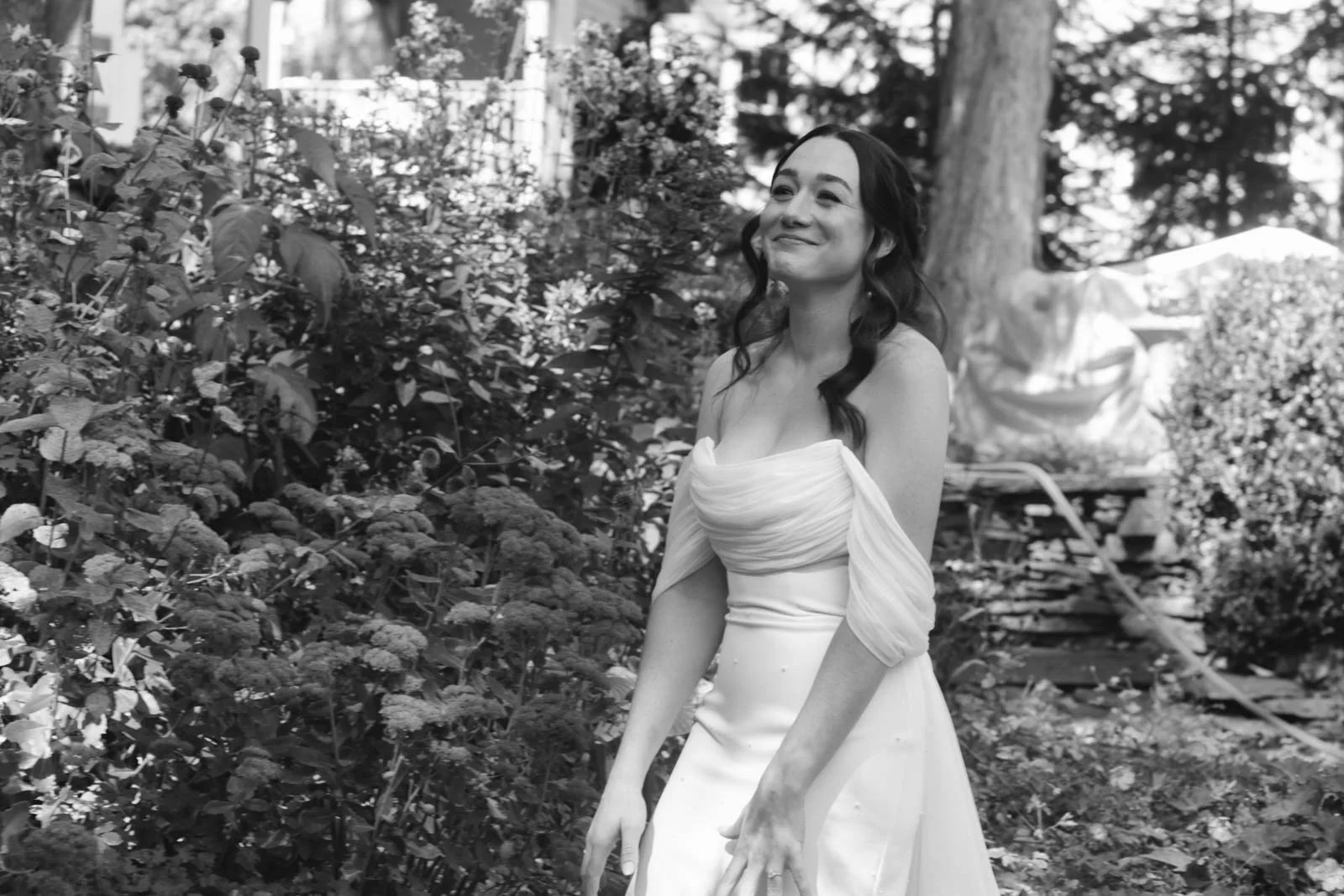A woman in a strapless wedding dress standing outdoors among plants and trees.