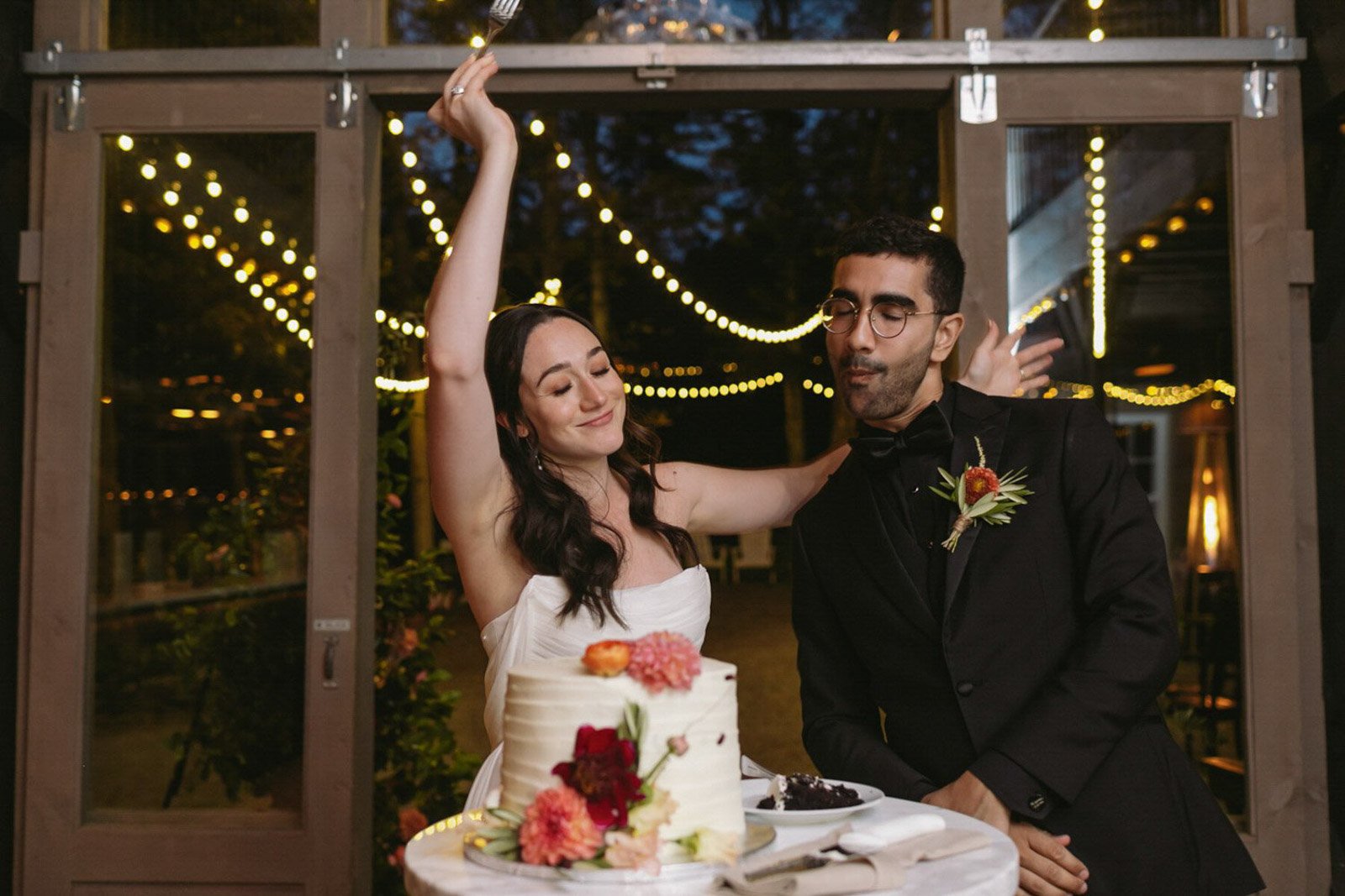 A bride and groom celebrate at their wedding reception, with the bride joyfully dancing and the groom standing nearby in a tuxedo with a boutonniere, a wedding cake decorated with flowers on the table, and string lights hanging overhead at night.