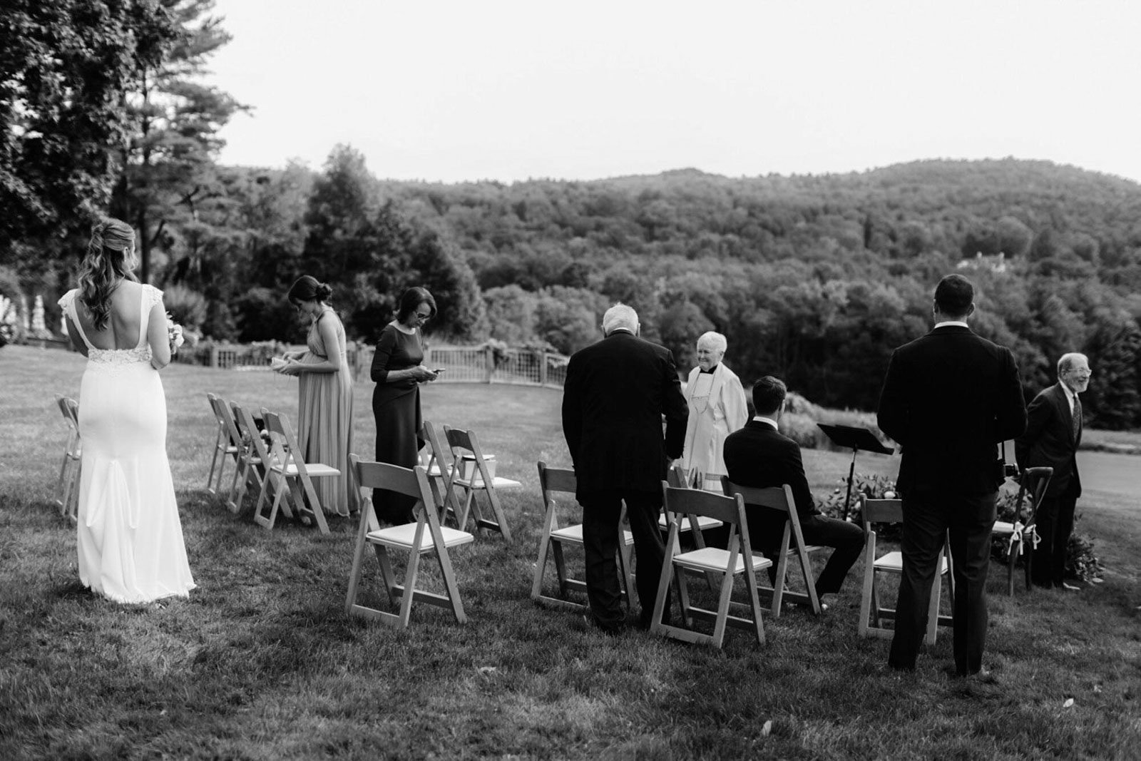 A black and white photo of a small outdoor wedding ceremony with guests standing and seated on the grass, with a scenic view of trees and hills in the background.