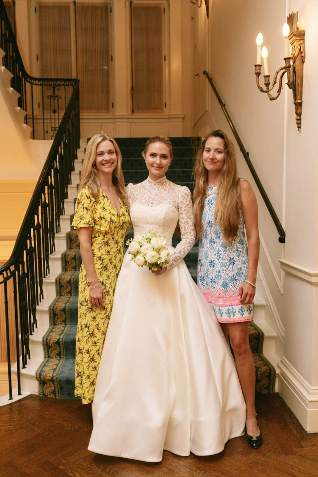 A bride in a white lace wedding dress holding a bouquet of white roses, standing between two women in colorful dresses, on a staircase inside a decorated venue.
