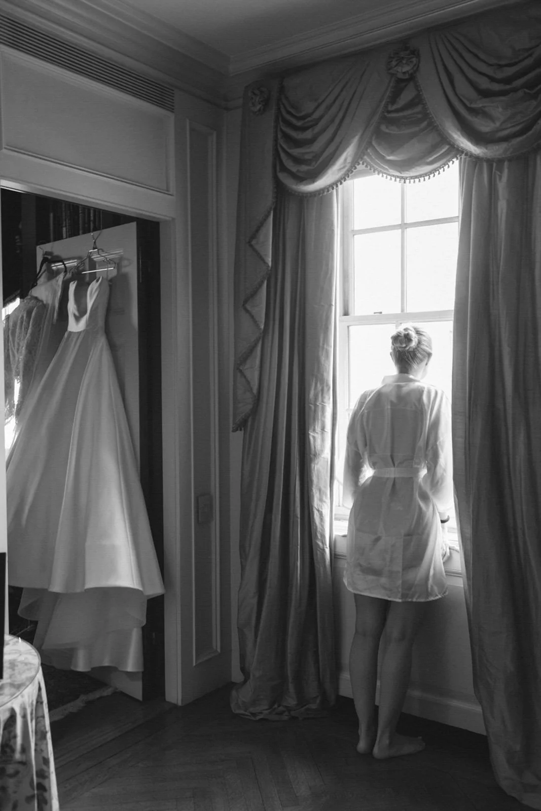 A woman stands in front of a window, looking outside, wearing a robe. Next to her, a dress is hanging on a closet door.