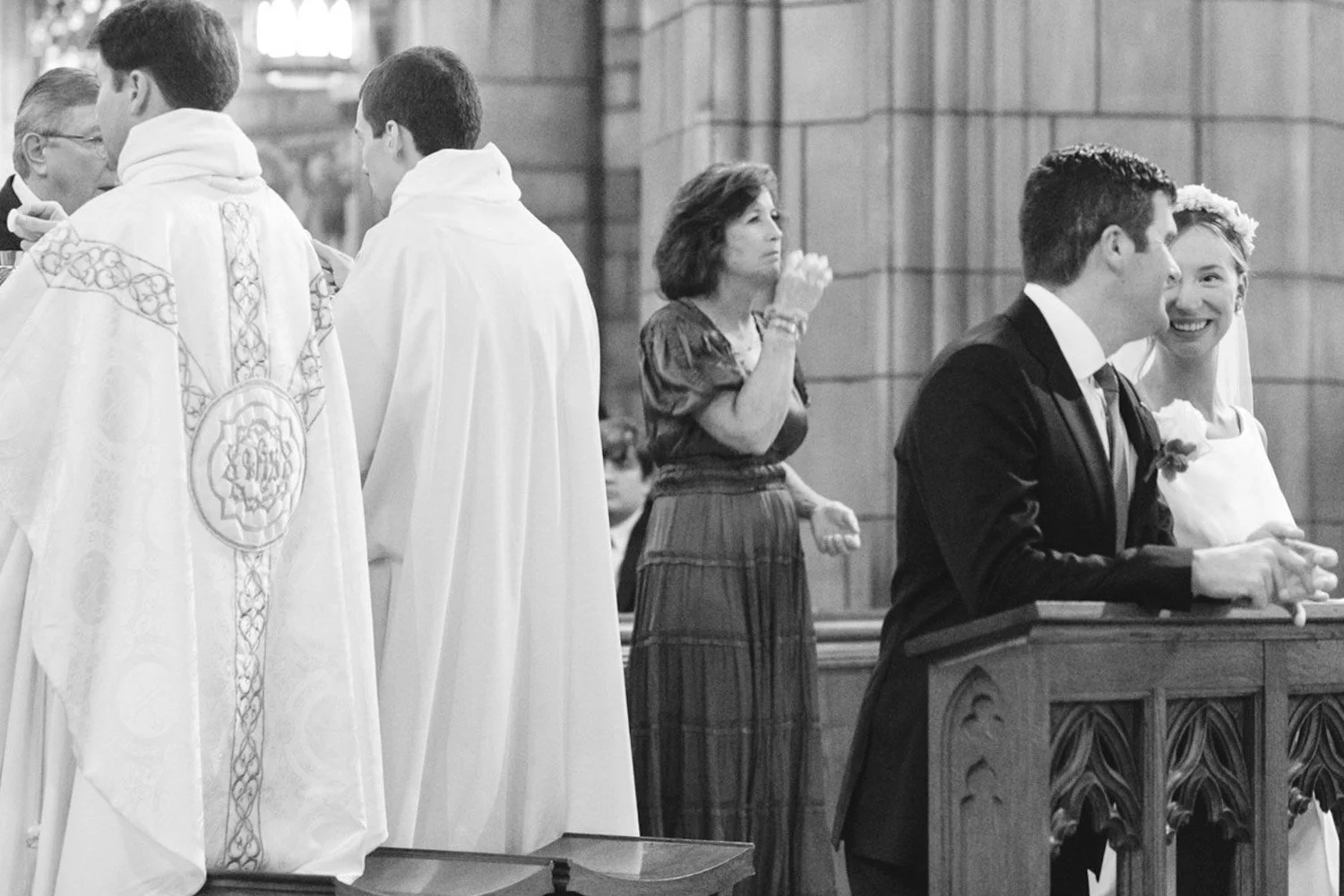 A black-and-white photo of a wedding ceremony inside a church, with the bride and groom standing at the altar, smiling, while a woman in the background appears to be praying or reflecting, and priests or officiants are present.
