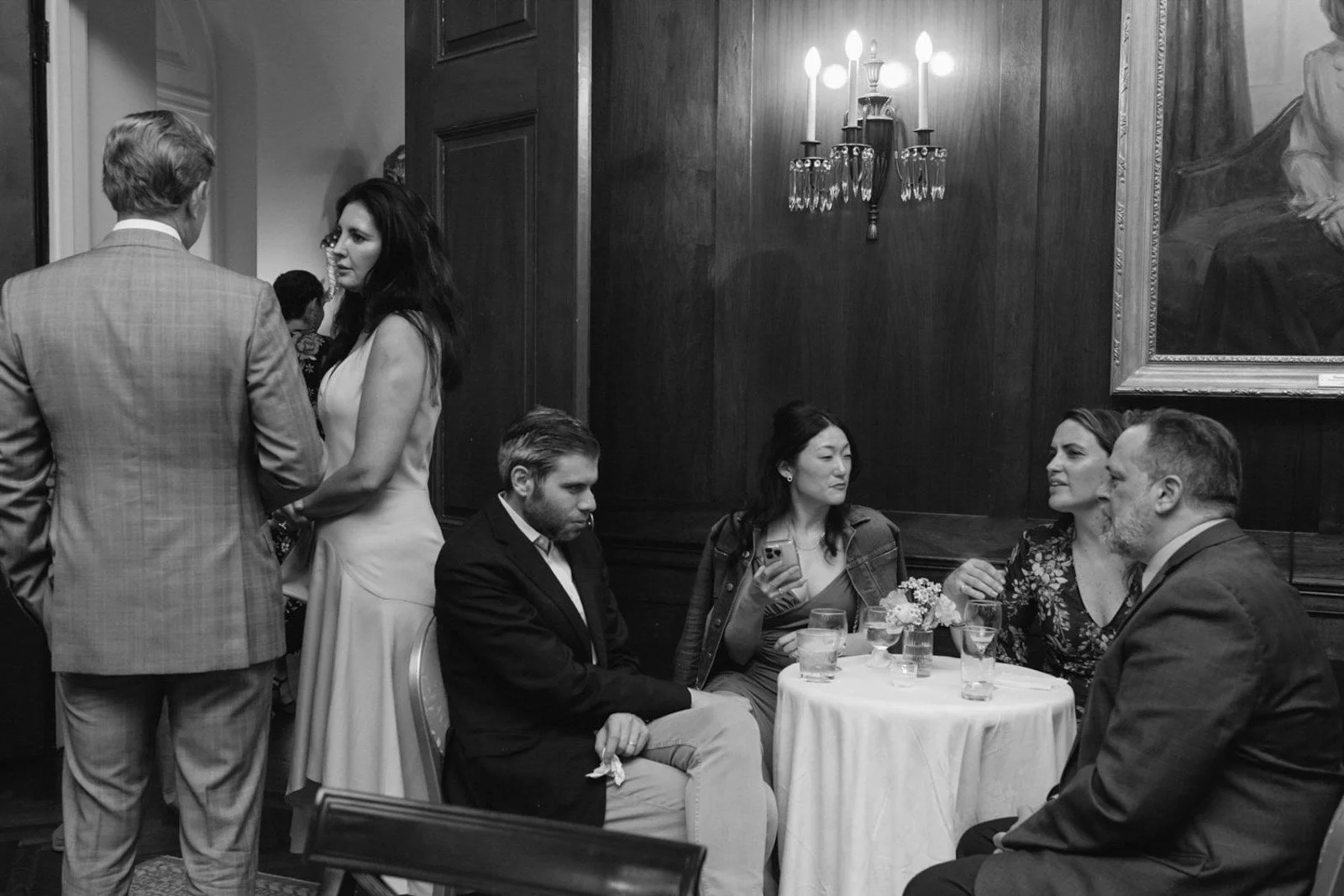 A group of six people socializing at a formal event, with three sitting around a table and three standing nearby, in a room with wood-paneled walls, a chandelier, and a framed painting.