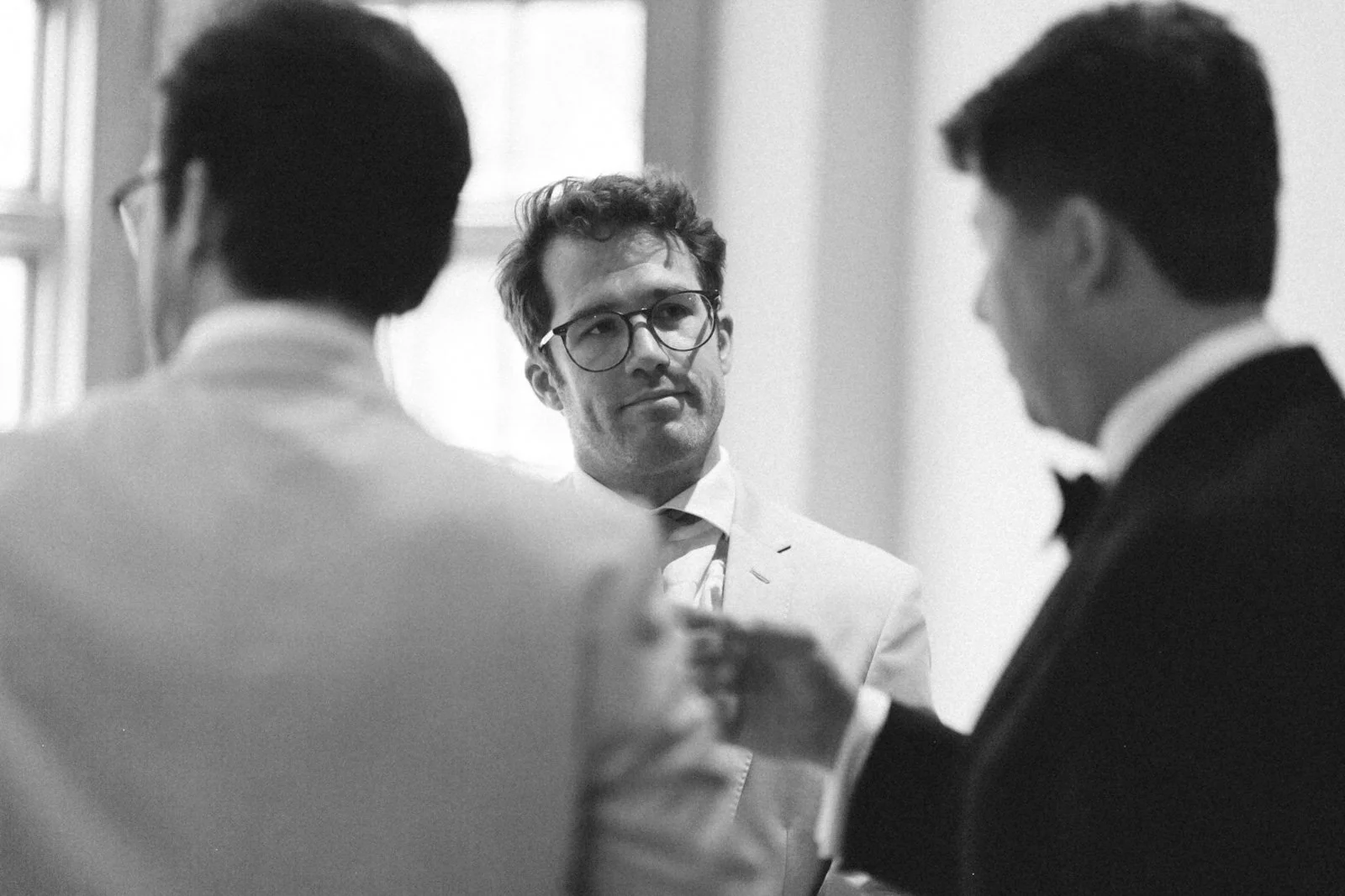 A black and white photo of three men engaged in conversation at a formal event, one man facing the camera and the other two with their backs to the camera, all wearing suits or tuxedos.