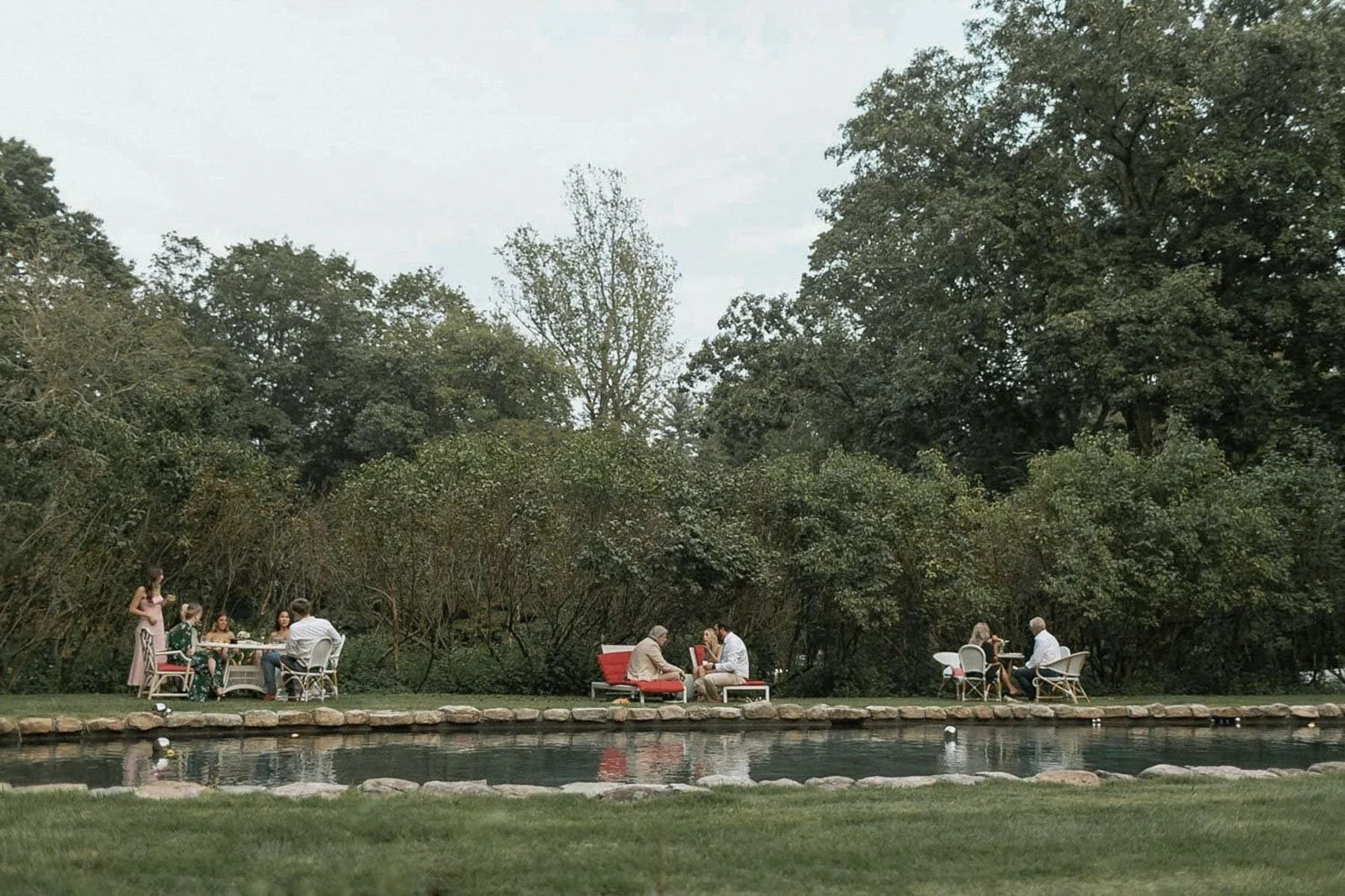 People enjoying an outdoor wedding by a pool with trees in the background.