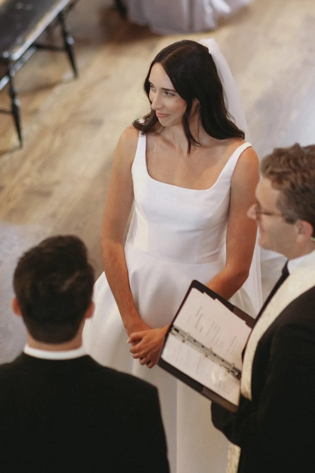 A woman in a white dress with dark hair and a veil, standing during a wedding ceremony surrounded by two men in suits, one holding a book or binder, in an indoor setting.