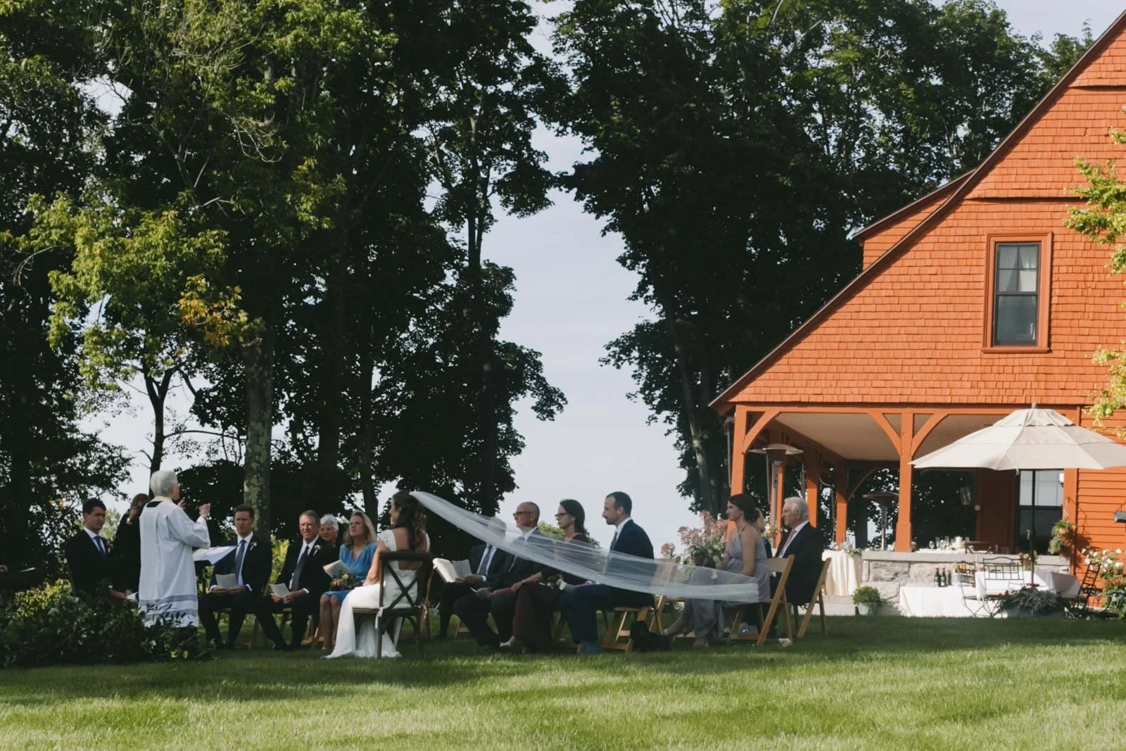 A wedding ceremony taking place outdoors on a lawn, with the officiant speaking to a seated bridal party, people in formal attire, next to a large red wooden house with an umbrella-covered table on the porch, and tall trees in the background.