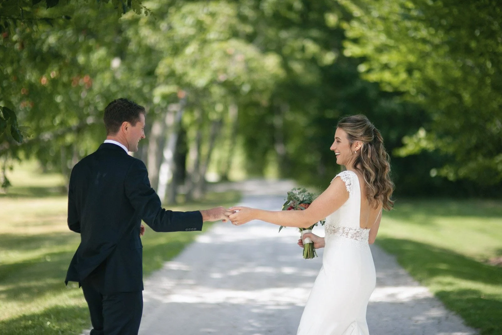 A bride and groom holding hands outside on a green, tree-lined path during a wedding, with the bride smiling and holding a bouquet.