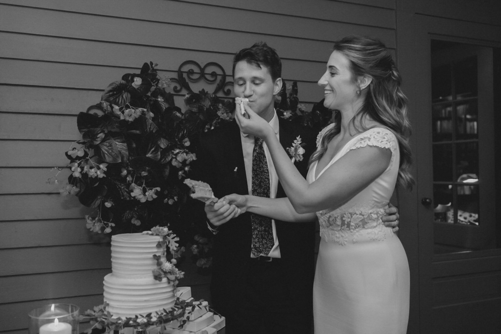 A bride and groom at their wedding cake cutting ceremony, with the bride feeding the groom a piece of cake, against a wooden wall backdrop.