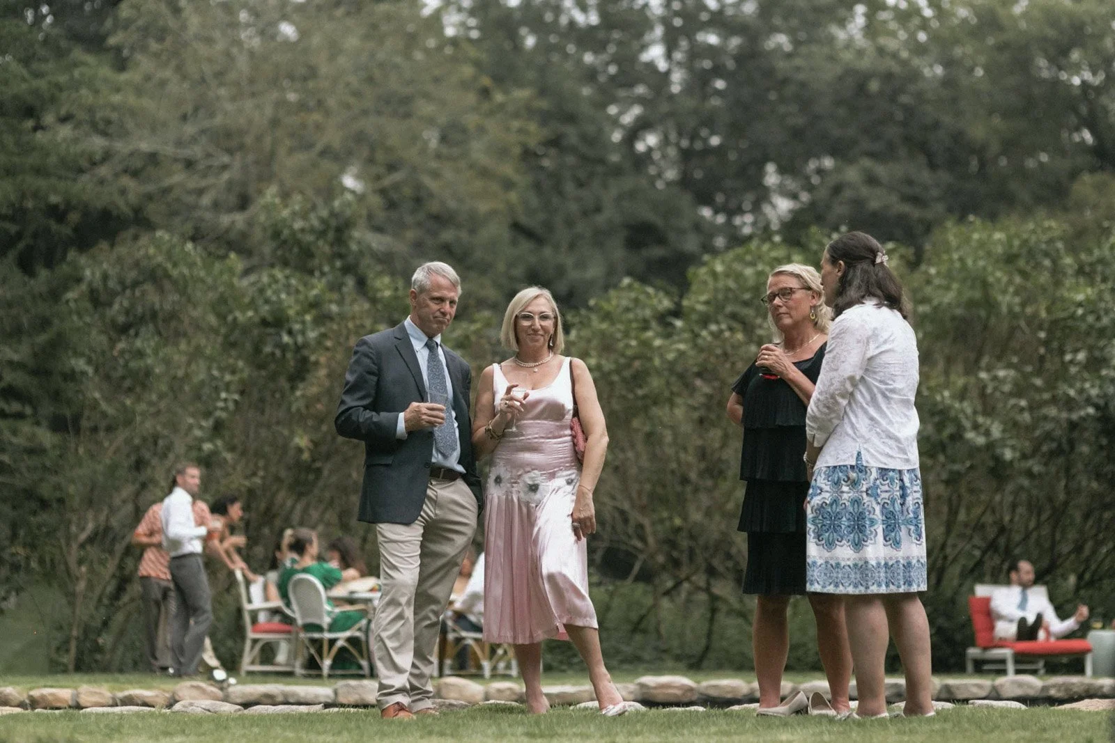 A group of four people standing outdoors on a lawn, engaged in conversation during an outdoor gathering with tables and chairs in the background.