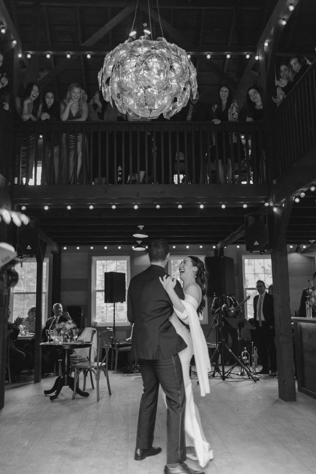A couple dancing at their wedding reception with guests watching from a second-floor balcony and seated at tables, interior of a rustic wedding venue.
