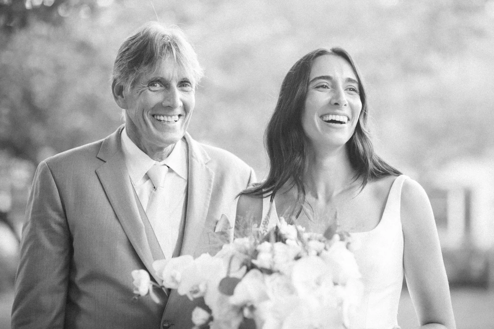 Black and white photo of a smiling older man in a suit and tie standing next to a young woman in a wedding dress holding a bouquet of flowers, outdoors with blurred trees in background.