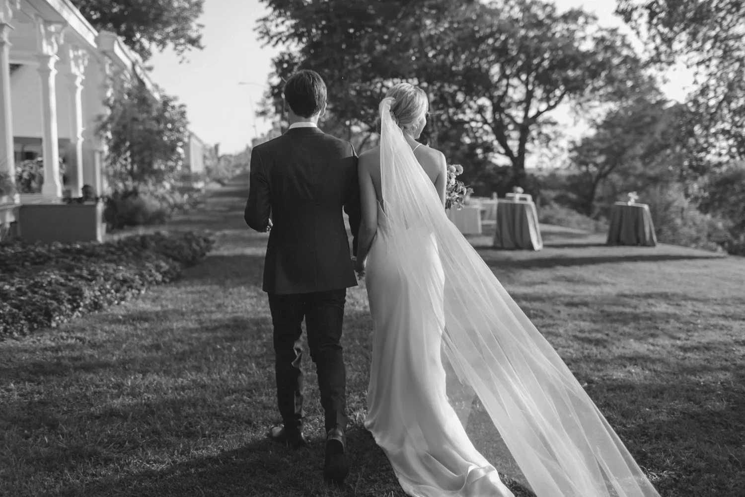 A bride and groom walking hand in hand outdoors during a wedding, with trees and wedding reception tables in the background, black and white photograph.