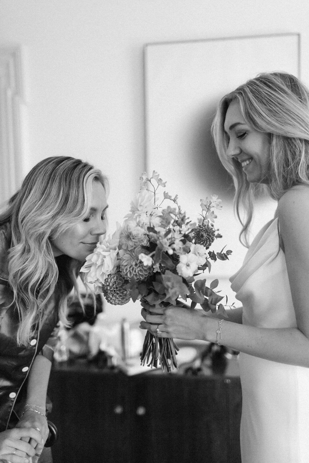 Two women, one holding a bouquet of flowers, are smiling at each other in a room.