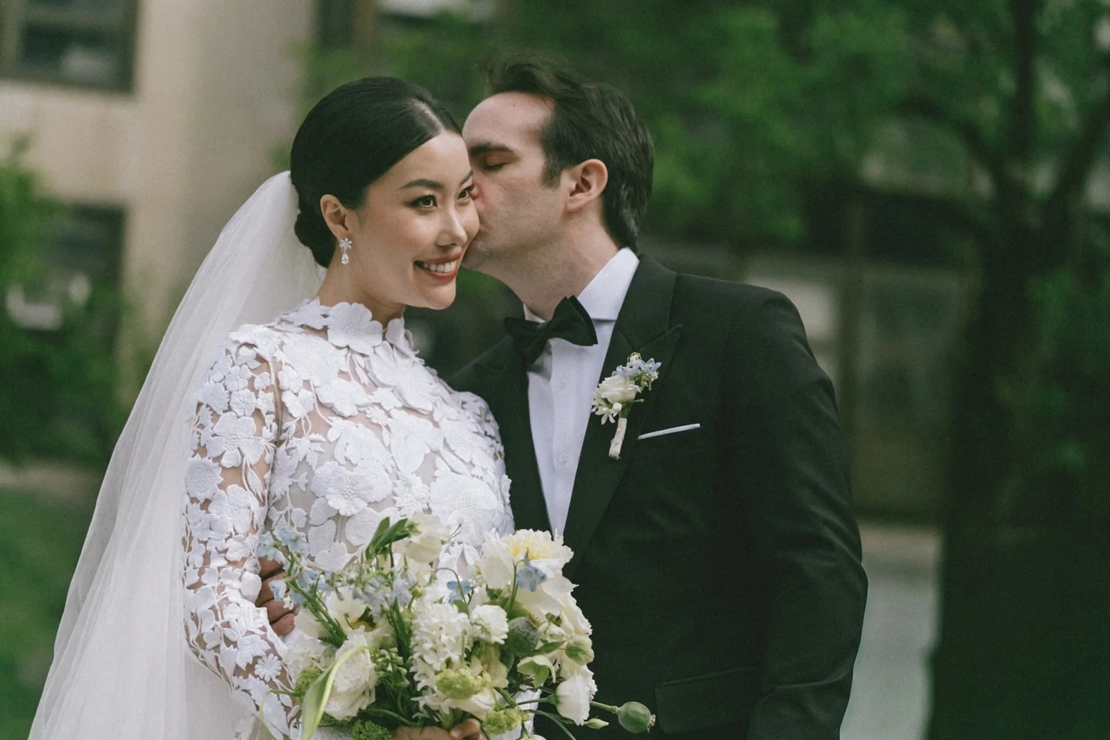 A bride and groom kissing during a wedding ceremony outdoors, with the bride holding a bouquet of white flowers and wearing a lace wedding dress, and the groom in a black tuxedo with a boutonniere.