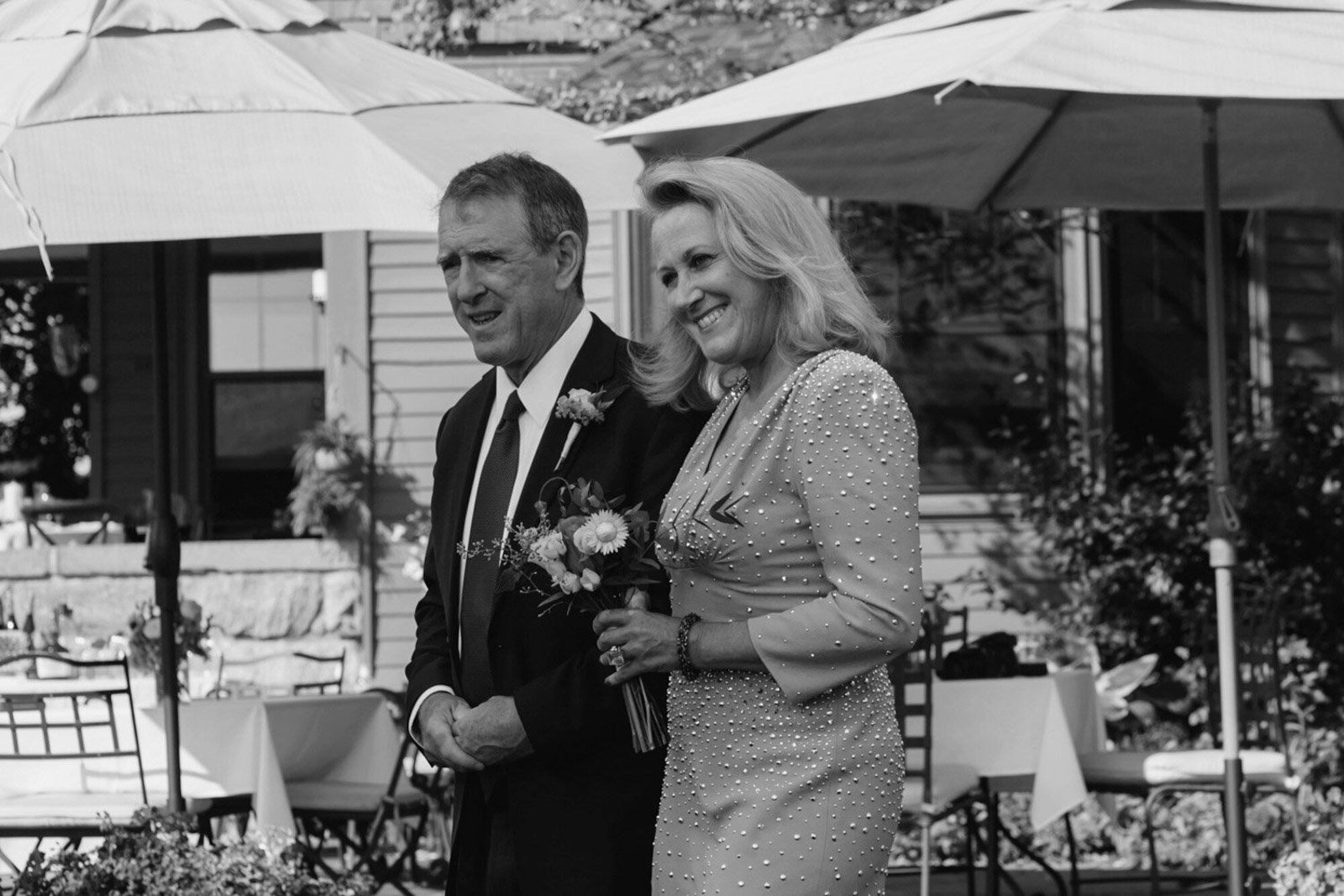 A black and white photo of a middle-aged man and woman walking together outdoors, with umbrellas overhead. The woman is smiling and holding a bouquet of flowers. The setting appears to be a garden or patio with tables and chairs.