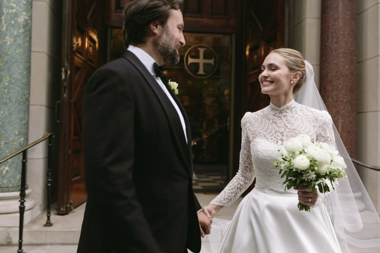 A bride and groom holding hands outside a church, smiling at each other. The bride is wearing a lace wedding dress and veil, holding a bouquet of white flowers. The groom is in a black tuxedo with a white shirt and black bow tie.