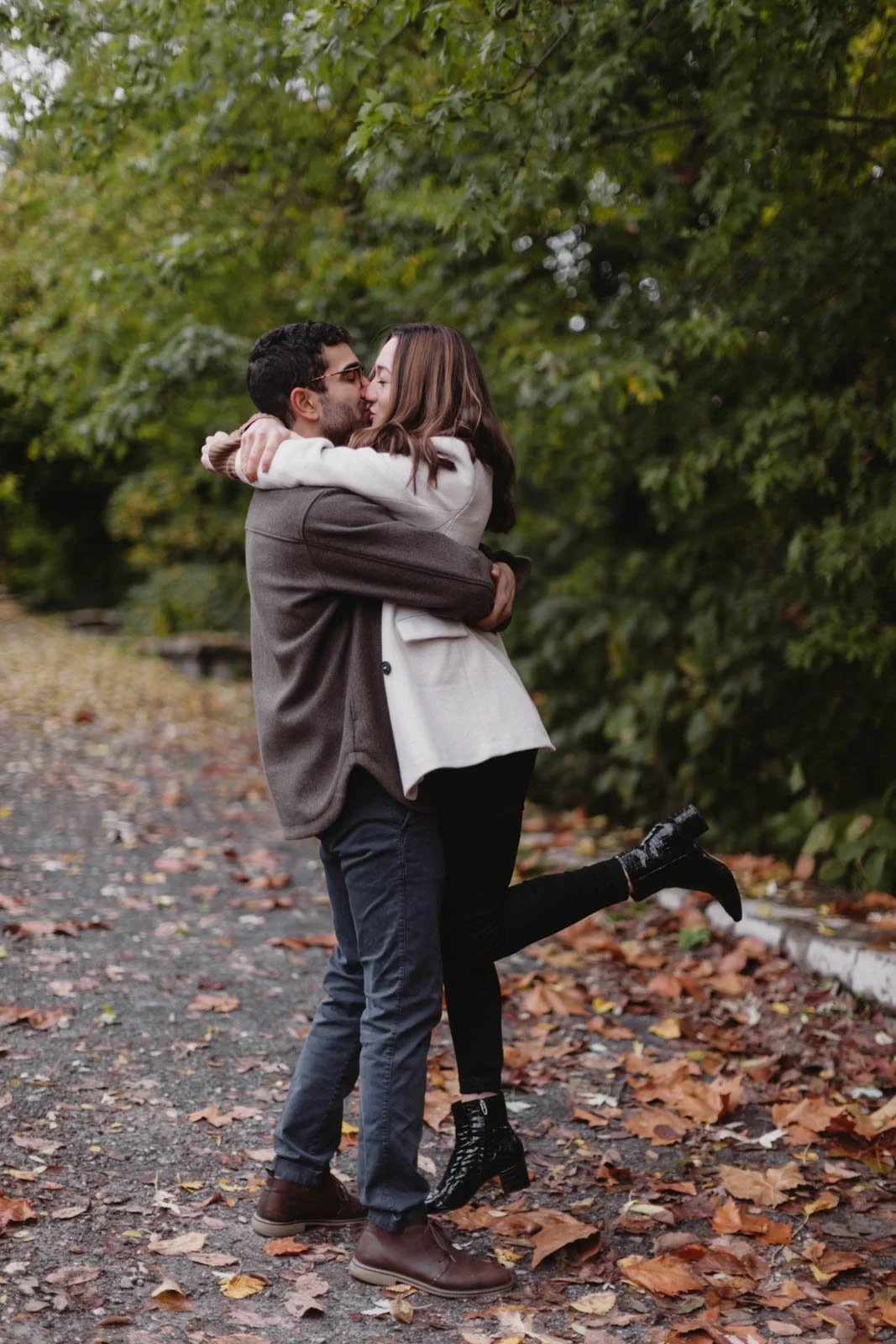 A young couple is hugging and kissing outdoors on a leaf-covered pathway during fall, with the woman lifting one leg and the man holding her.