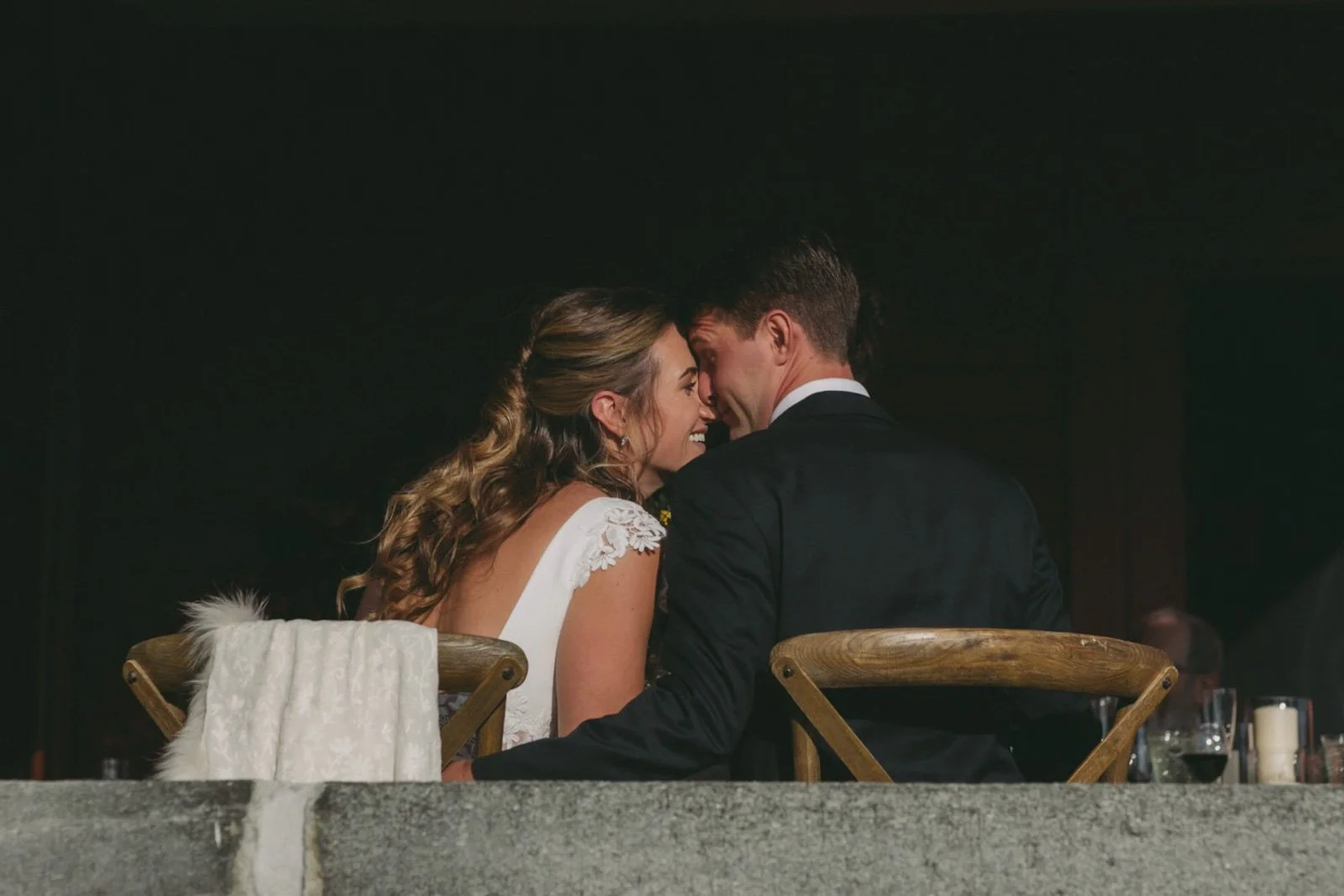A bride and groom sitting close together at a wedding reception, touching noses and smiling.