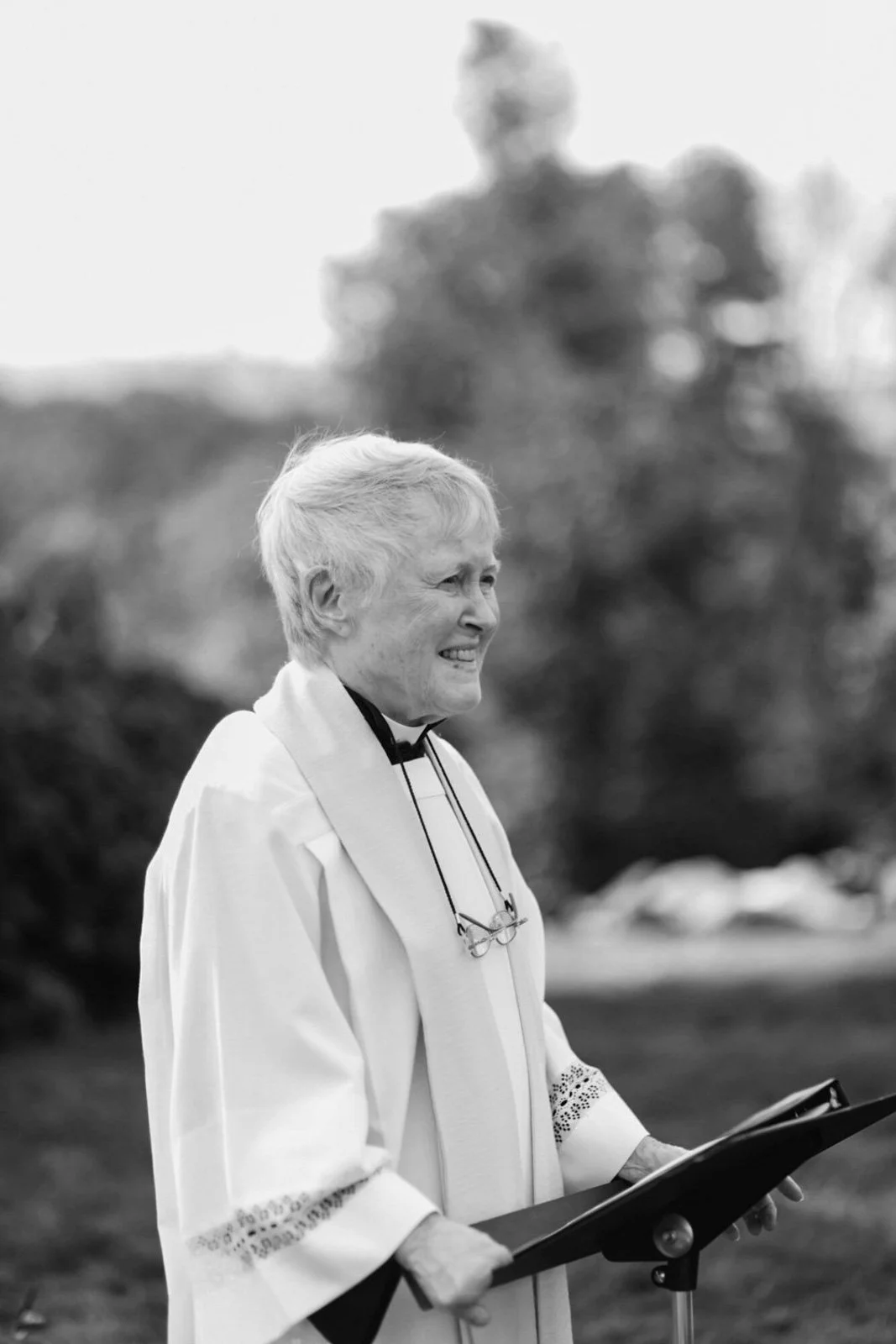 An elderly woman dressed as a clergy member standing outdoors, holding a book or folder, with a blurred background of trees.
