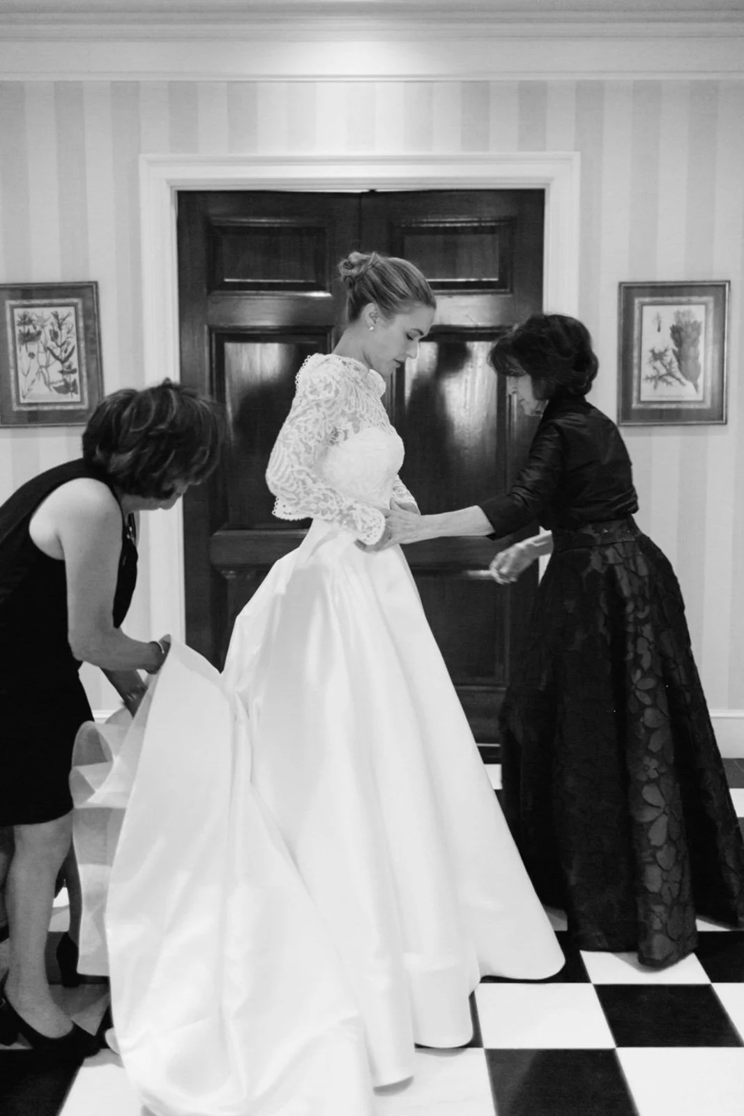 A woman in a wedding dress is being assisted by two others in a room with striped wallpaper and framed artwork, a wooden door in the background, and a checkered floor.