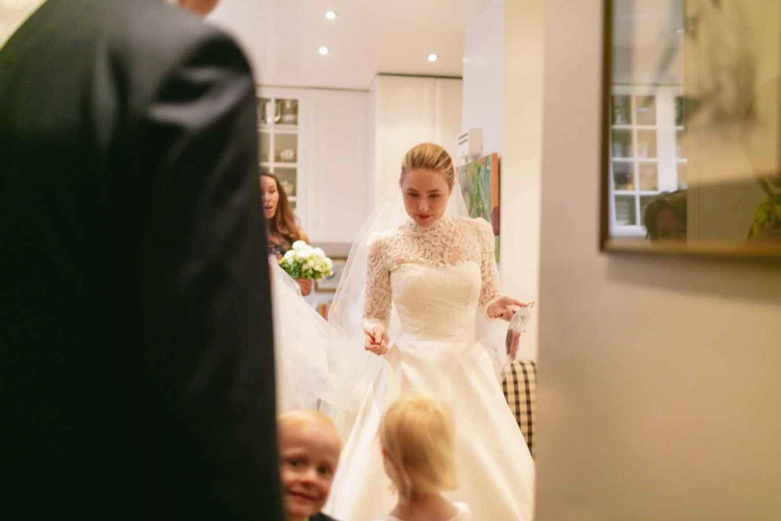 A bride in a white wedding dress with lace sleeves and a veil, holding her dress while standing in a room with children and a woman holding a bouquet of flowers in the background.