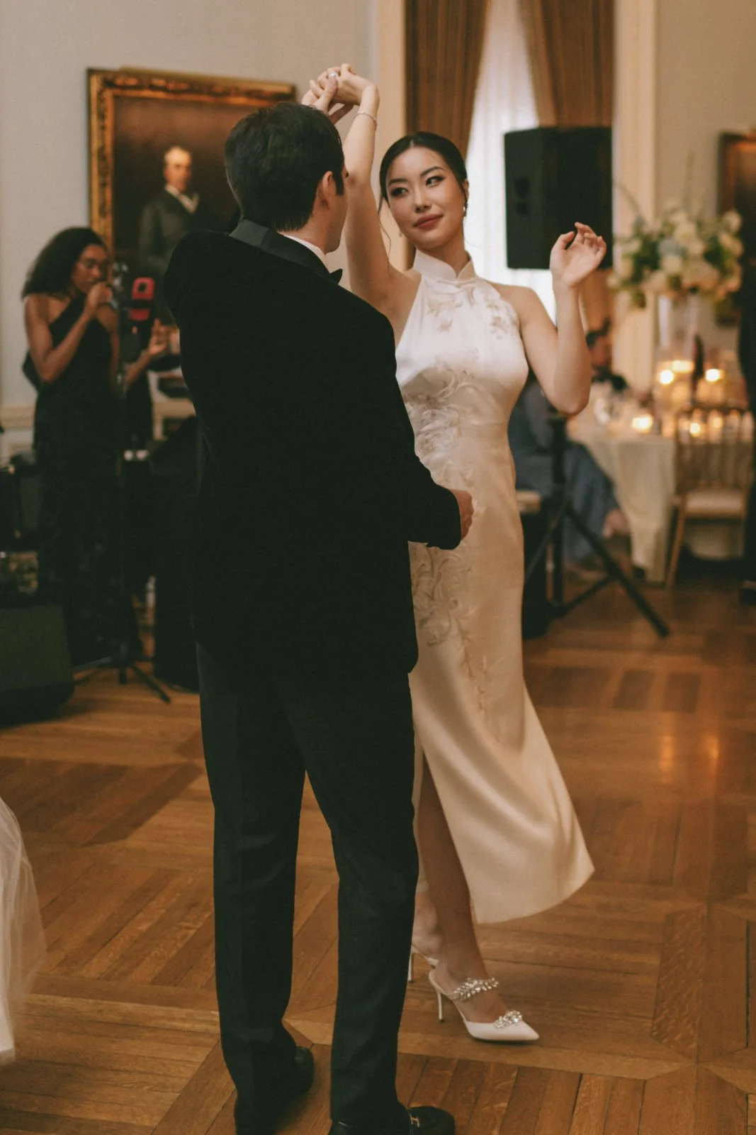 A couple is dancing at a formal event indoors, with a woman in a black dress taking a photo in the background.