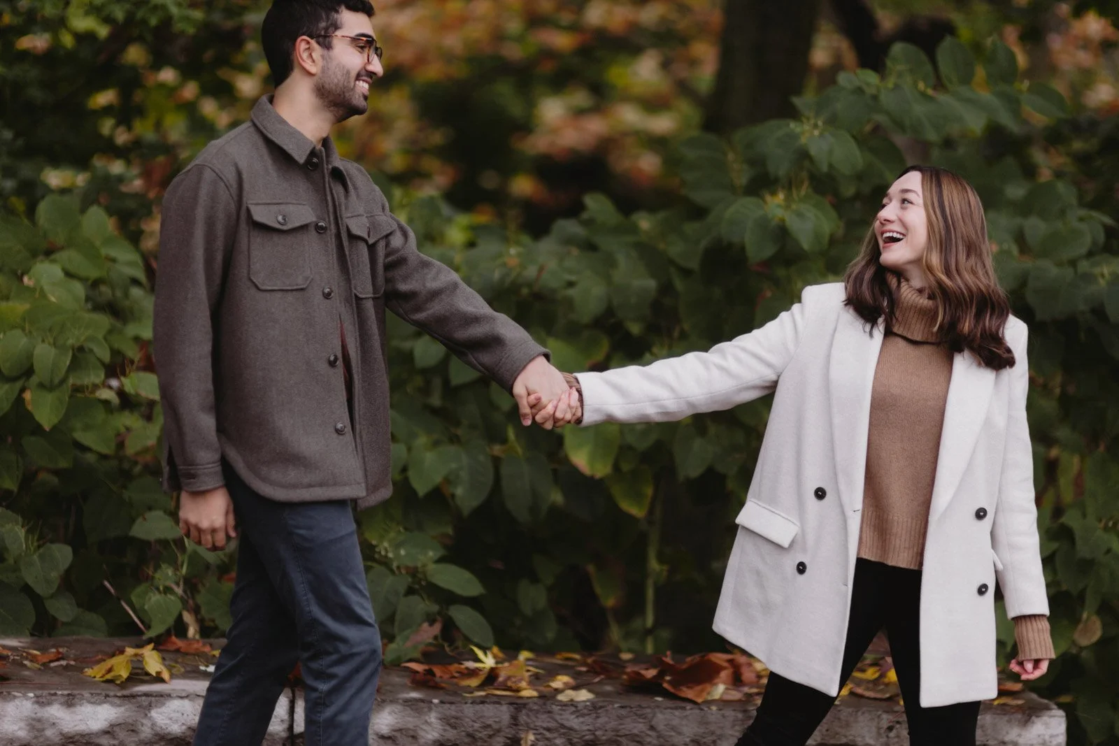 A man and woman holding hands outdoors, smiling at each other with a background of green leaves and autumn foliage.