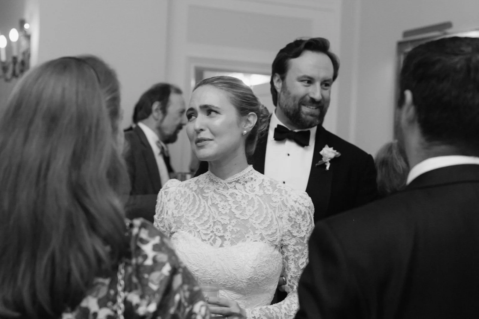 Black and white photo of a wedding reception, featuring a woman in a lace wedding dress listening to a man in a tuxedo with a bow tie, who is smiling. Several other guests are in the background having conversations.