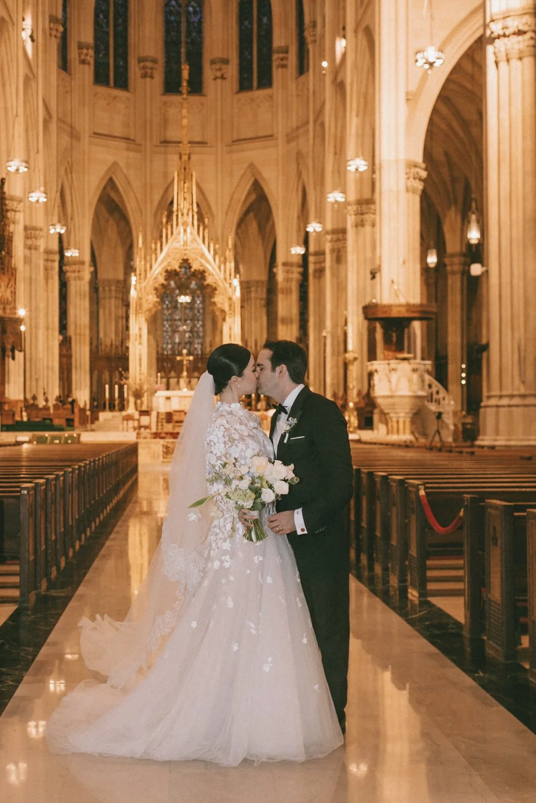 A bride and groom kissing inside a grand, ornate church. The bride is wearing a white lace wedding gown with a long veil, holding a bouquet of white roses. The groom is dressed in a black tuxedo. The church features high vaulted ceilings, stained gla