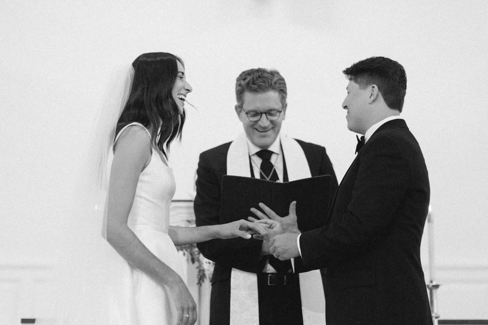A black-and-white photo of a wedding ceremony showing a bride and groom exchanging rings, with a officiant standing between them, all smiling.