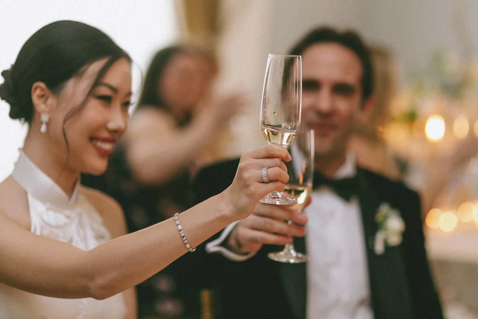 A woman in a white dress smiling and holding a champagne flute, standing next to a man in a tuxedo at a celebration or wedding reception.
