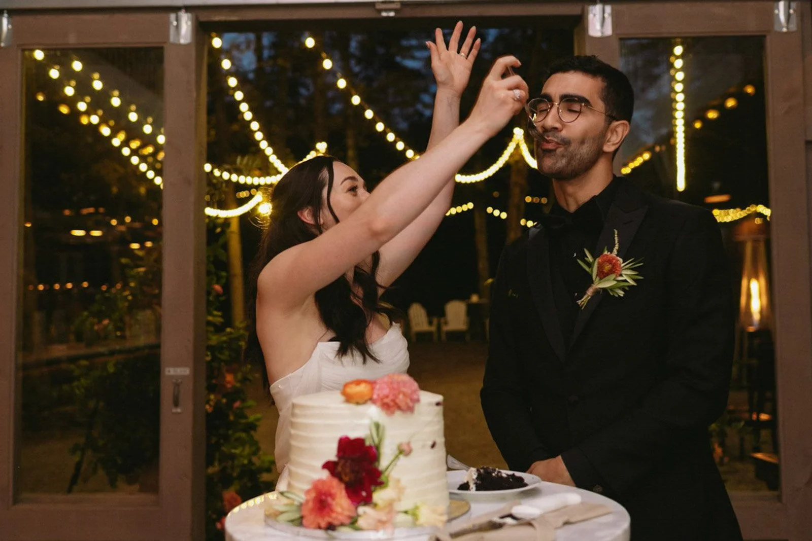 A woman in a white wedding dress and a man in a black suit are celebrating at their wedding reception, with string lights hanging in the background. The woman is feeding cake to the man, who is smiling with closed eyes. A wedding cake decorated with 