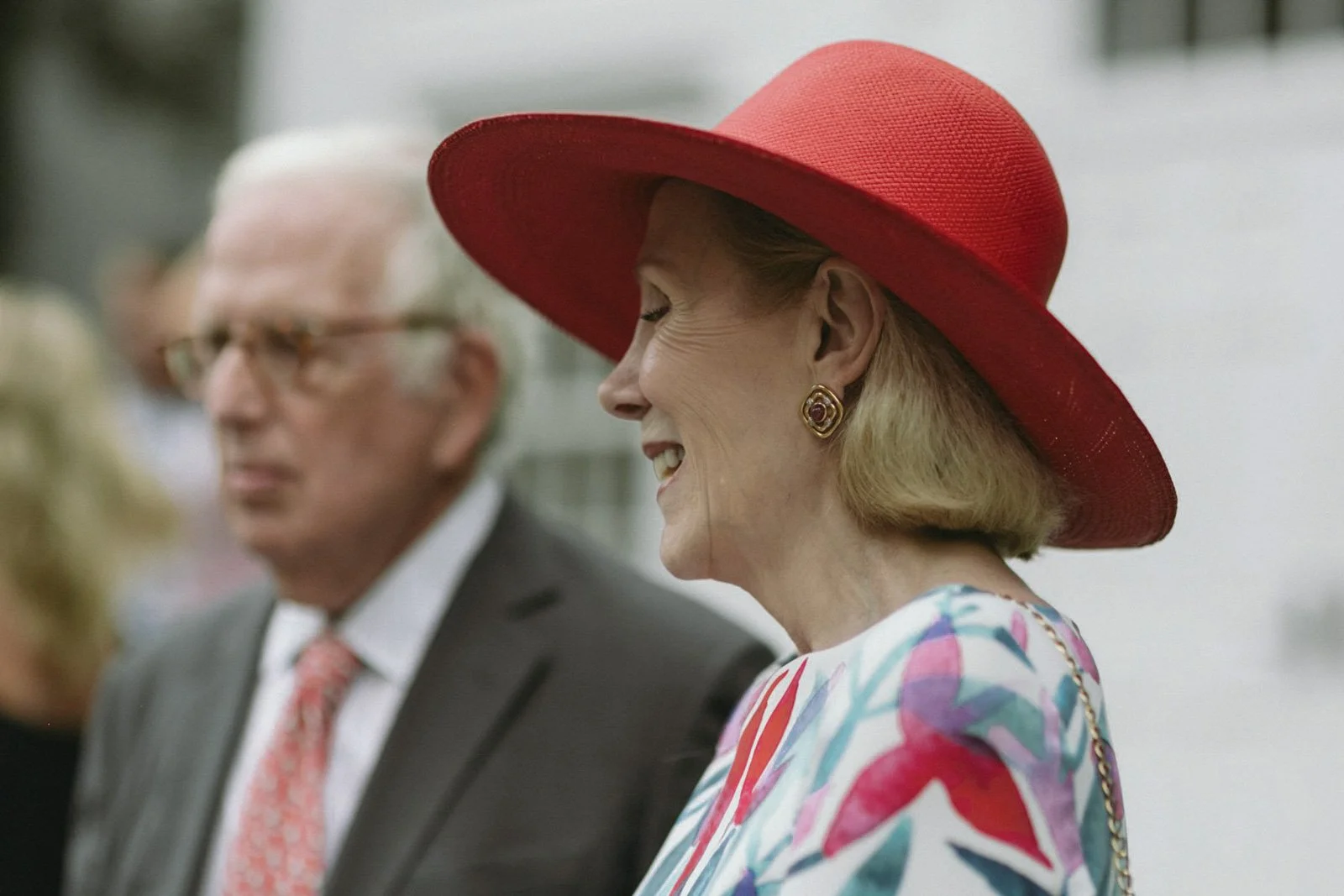 A woman wearing a large red hat and colorful dress, smiling, with an older man in a suit and tie in the background.