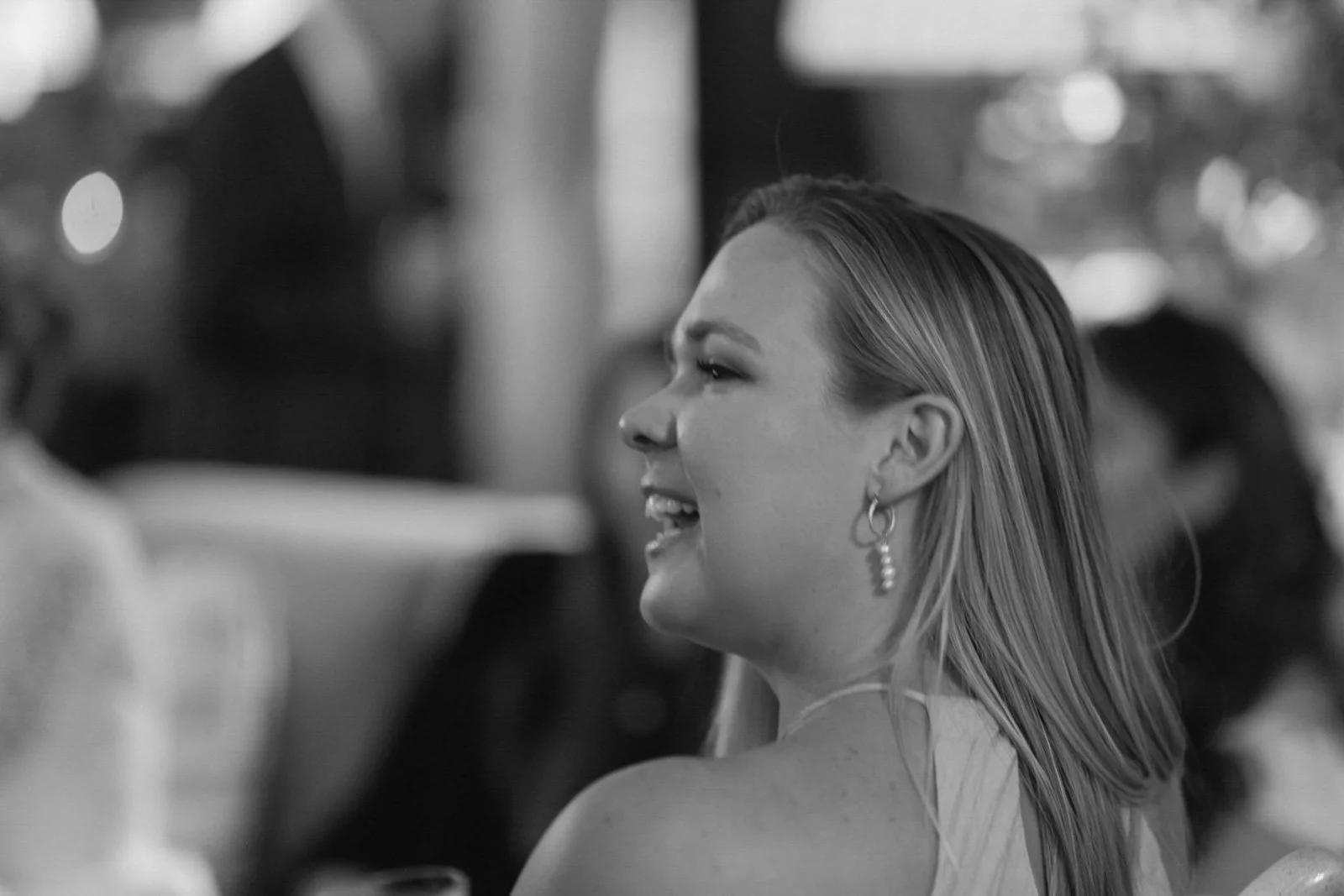 A woman with light-colored hair, smiling and wearing earrings, appears to be in a social setting with blurred background figures.