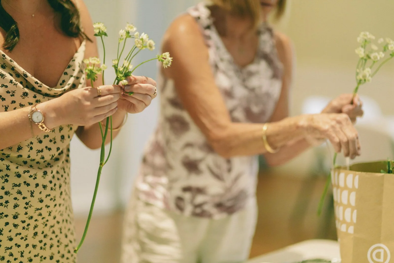 Two women holding flowers while preparing floral arrangements indoors.