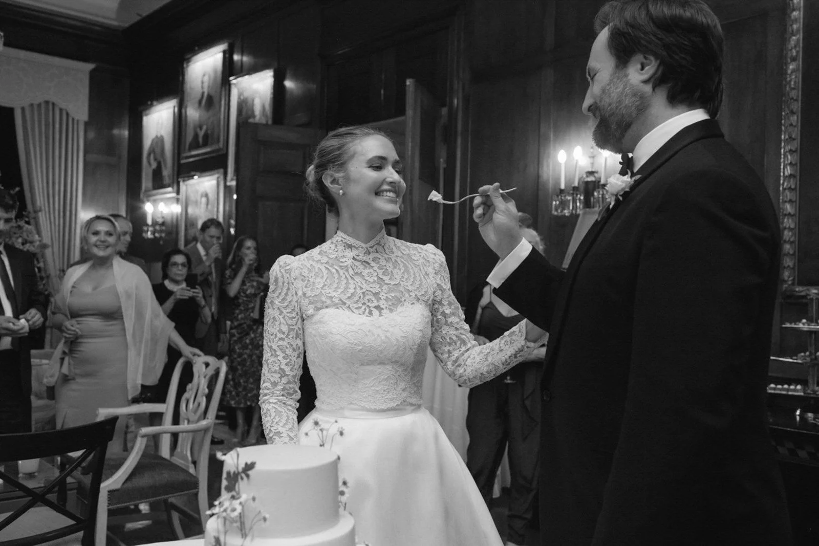 A black and white photo of a wedding reception where a bride and groom share a cake. The bride is smiling and looking at the groom, who is feeding her cake with a fork. Guests watch and smile in the background.