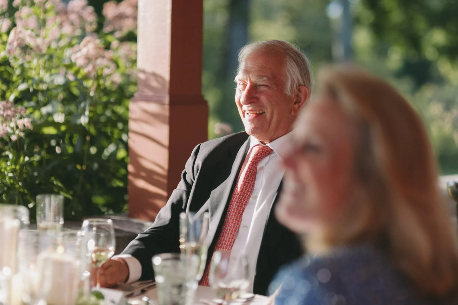 Older man in a suit and red tie sitting outdoors, smiling and enjoying a sunny day, with a woman partially visible in the foreground.