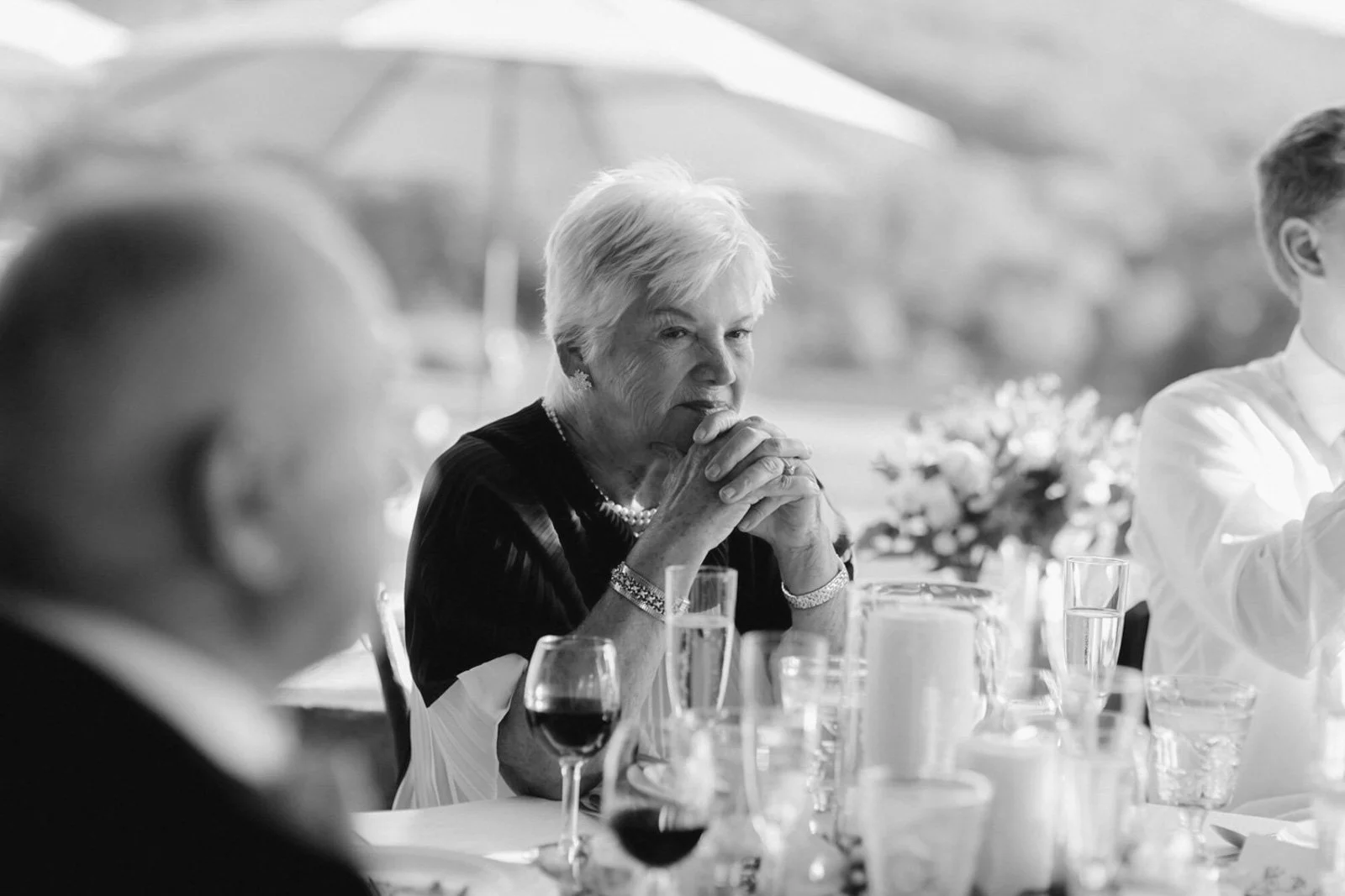 An elderly woman with short hair sitting at a table, clasping her hands near her mouth, with a glass of red wine in front of her, during a social gathering.