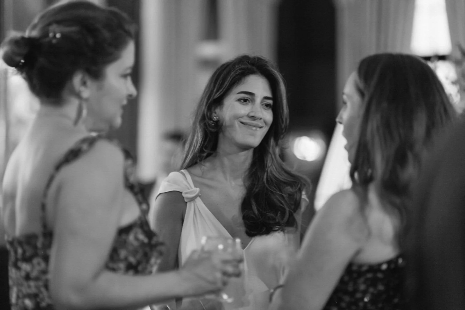Three women are having a conversation at a social gathering. The woman in the middle, with long dark hair, is smiling and holding a drink. The other two women, blurred, are facing her, and one is holding a glass.