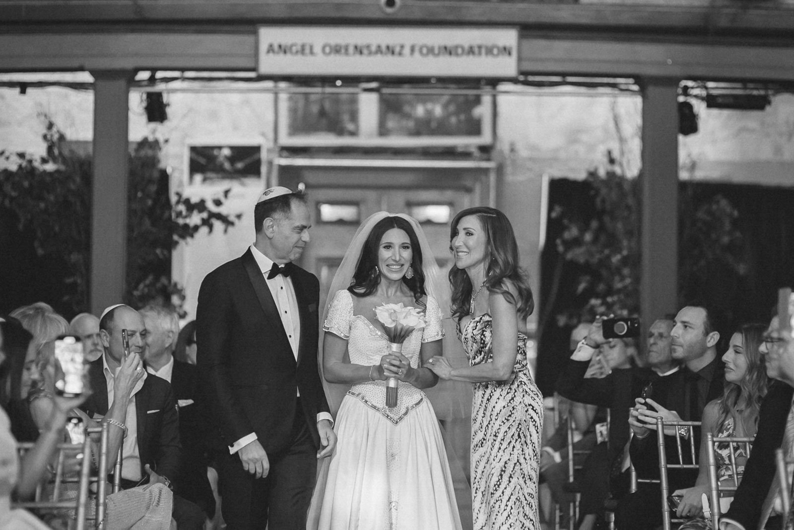 Wedding ceremony with a bride holding flowers, a groom in a tuxedo, and a woman in a patterned dress, all under a sign that reads 'Angel Orensanz Foundation', with guests seated and taking photos.