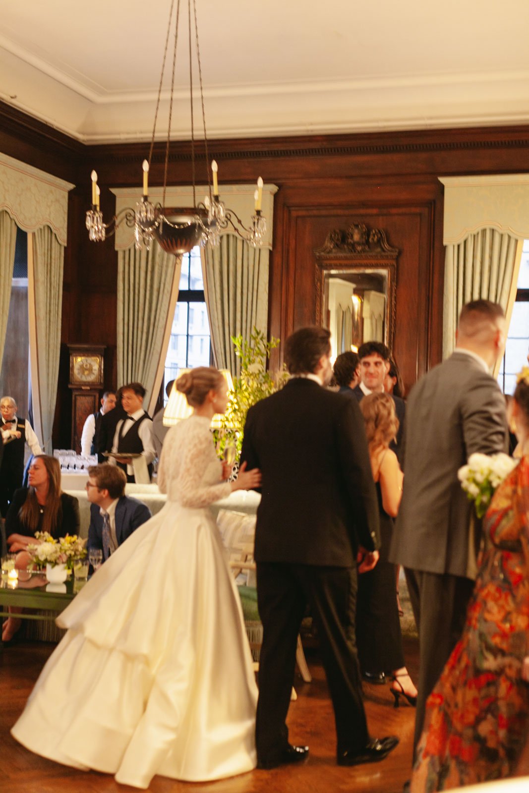 A bride in a white wedding dress stands next to a man in a black tuxedo at a formal event, surrounded by other well-dressed guests in an elegant, wood-paneled room with large windows, curtains, and a chandelier.