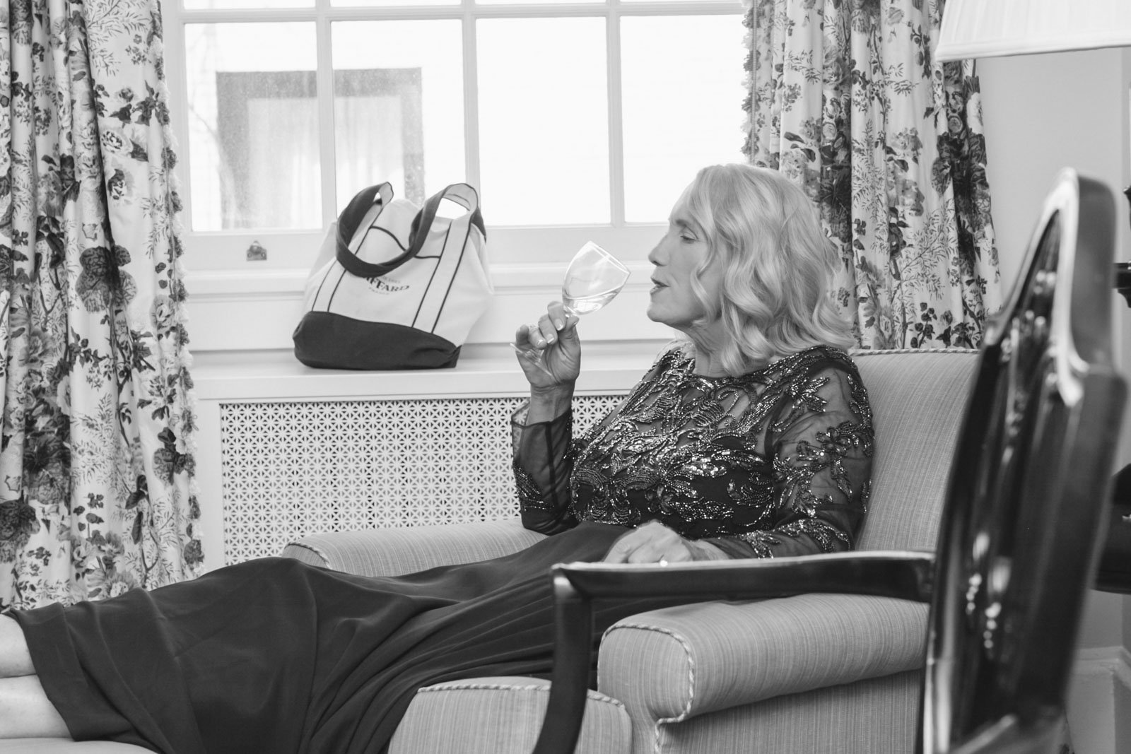 A woman with wavy blonde hair sitting on a sofa, sipping from a wine glass, wearing a dark, embellished dress in a room with floral curtains and a window.
