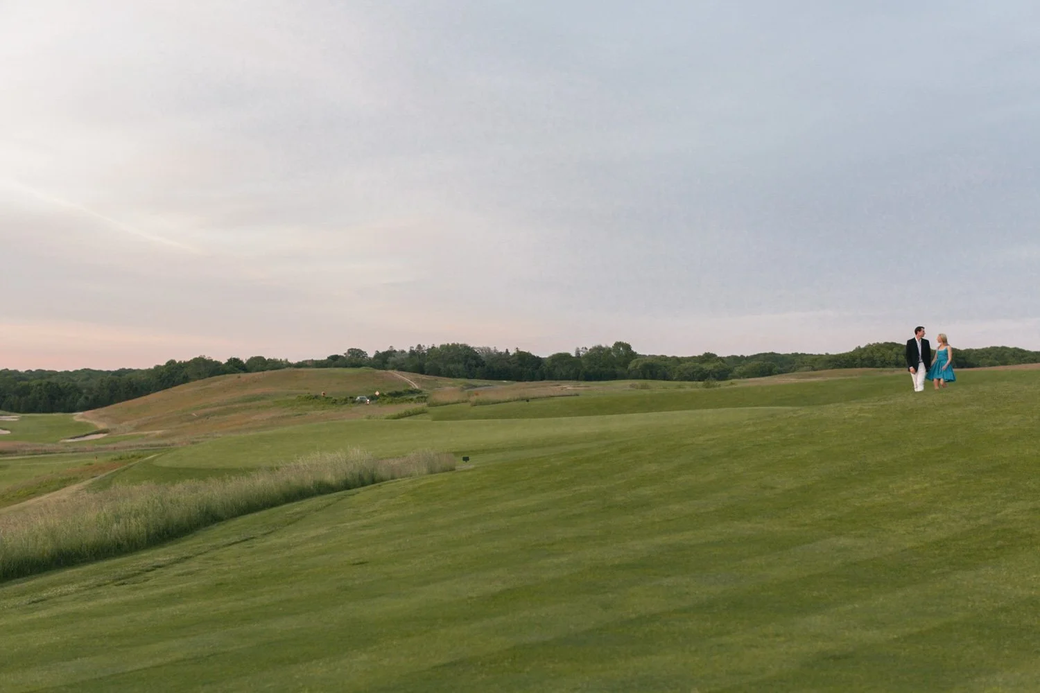 A man in a black suit and a woman in a blue dress walking at The Shinnecock Golf Club, at sunset.