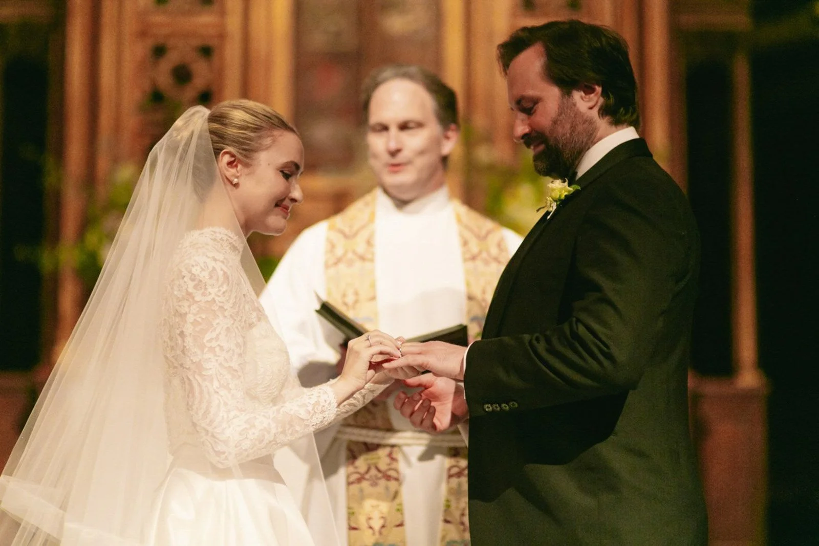A bride and groom exchanging wedding rings during their ceremony in a church, with an officiant standing behind them.
