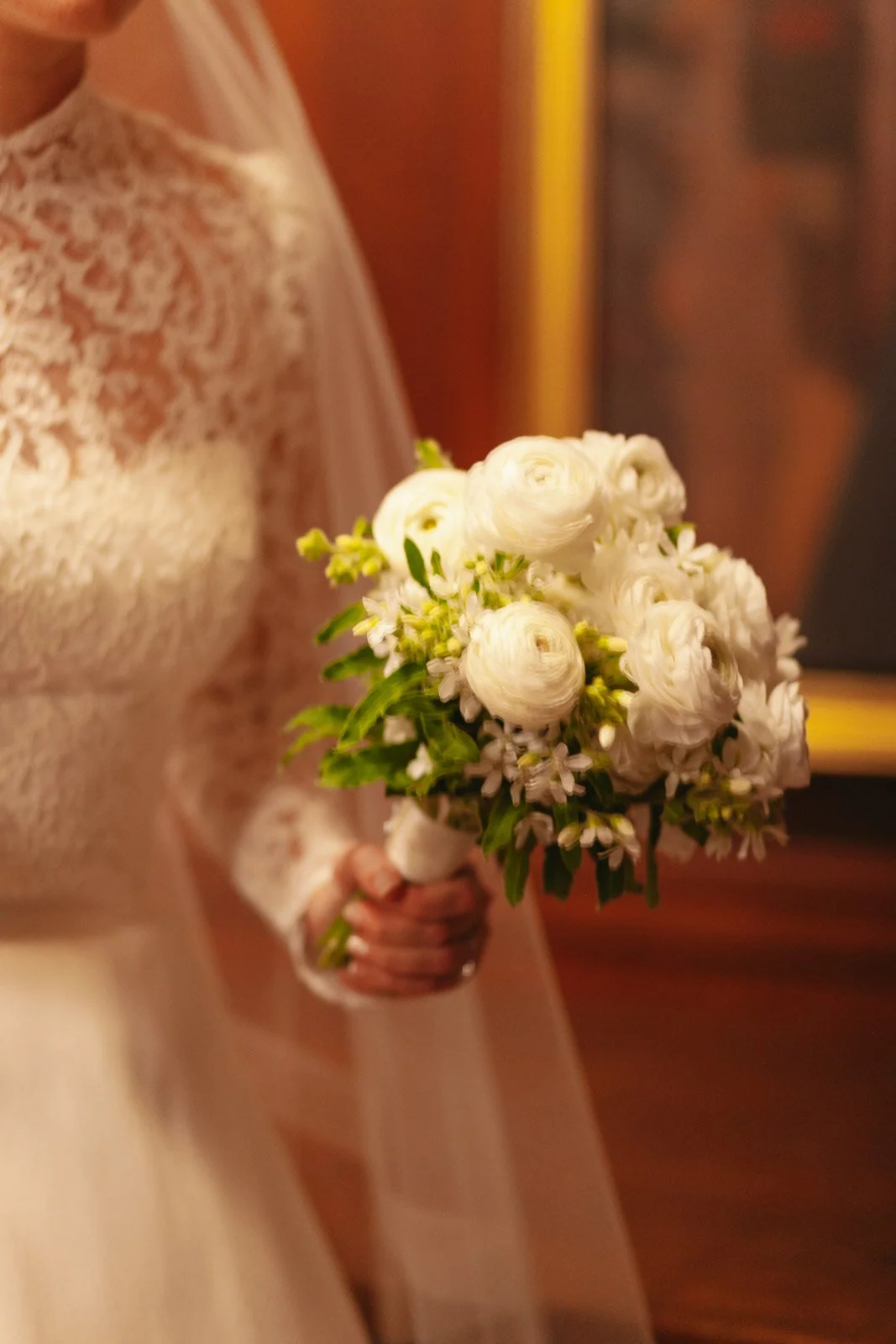 A bride in a lace wedding dress holding a bouquet of white flowers, including ranunculus and small daisies.